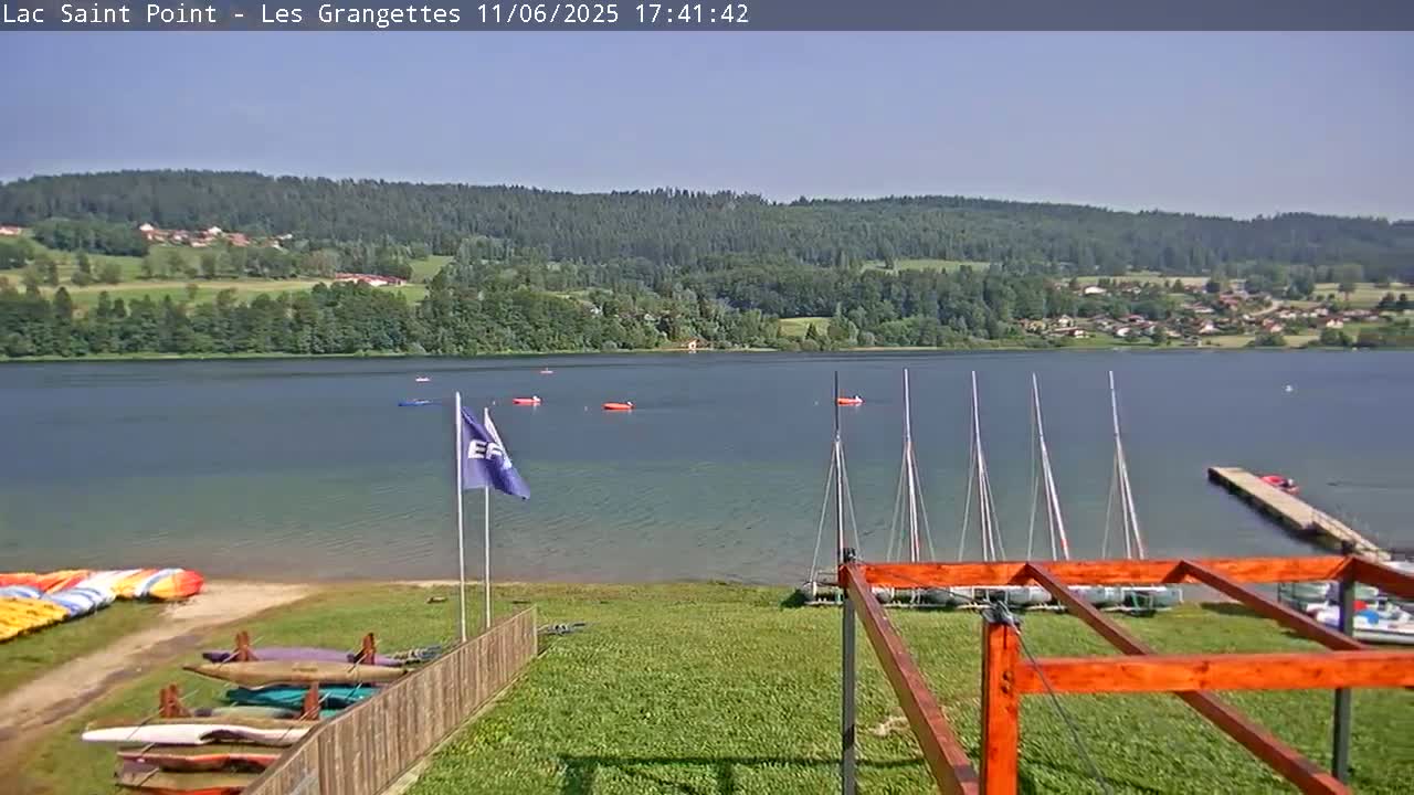 A calm lake under a clear blue sky is surrounded by green hills and trees, with sailboats on the water and various water sports equipment stored on the shore.