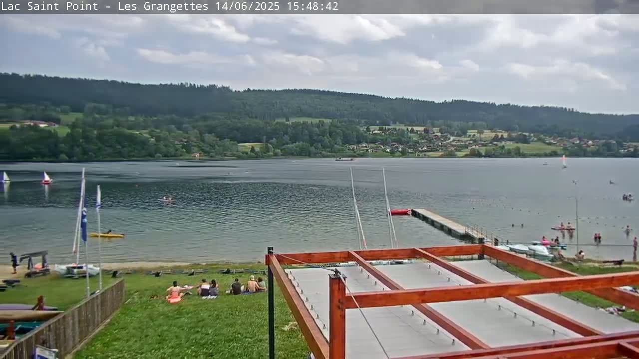 A partly cloudy day at a lake with people swimming, boating, and sunbathing on the shore, with a wooden structure in the foreground.