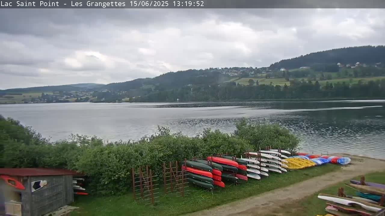 A calm lake under a cloudy sky is viewed from a lakeside location, where numerous kayaks and canoes are stored on racks near a small shed.