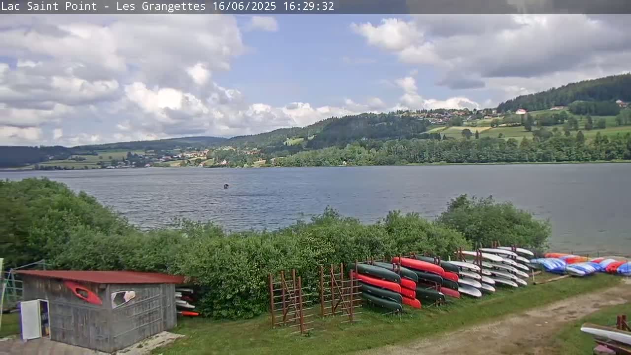 A mostly cloudy day reveals a calm lake surrounded by green hills and trees, with numerous canoes stored on racks near a small shed.