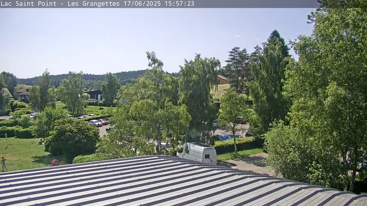 A metal roof overlooks a sunny parking lot surrounded by lush green trees and a grassy area, with a hilly landscape in the background.