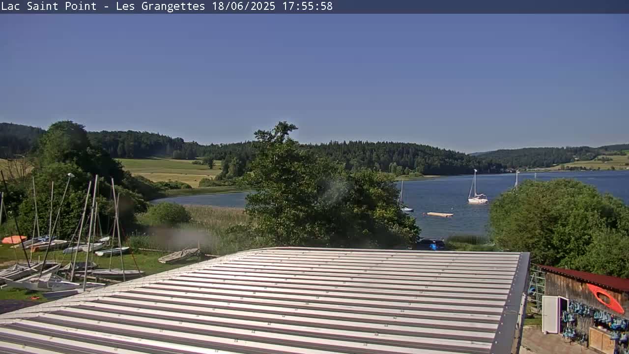A corrugated metal roof overlooks a calm lake under a clear blue sky, with sailboats on the water and several sailboats docked on the shore near a building.