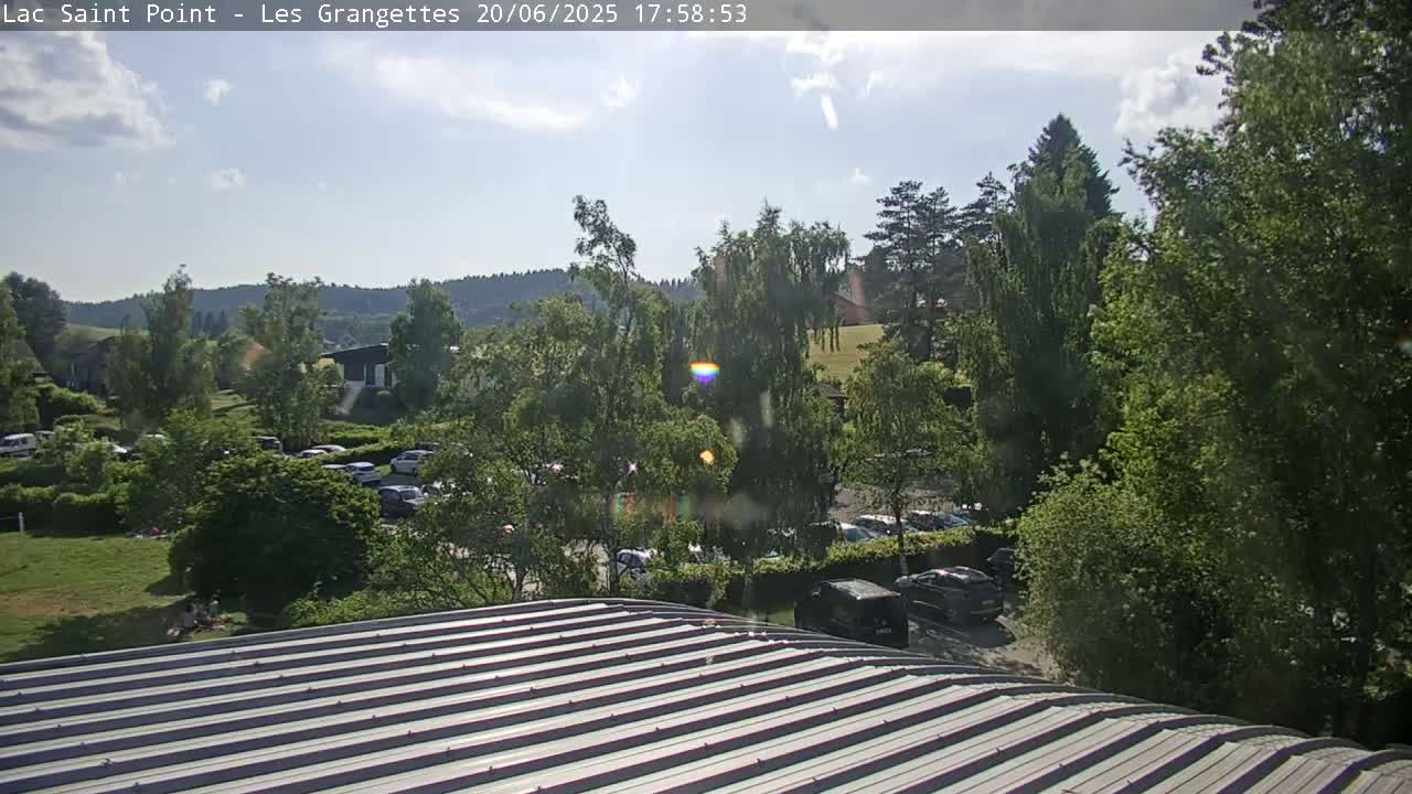 A corrugated metal roof is in the foreground, overlooking a parking lot surrounded by lush green trees and hills under a partly cloudy sky.