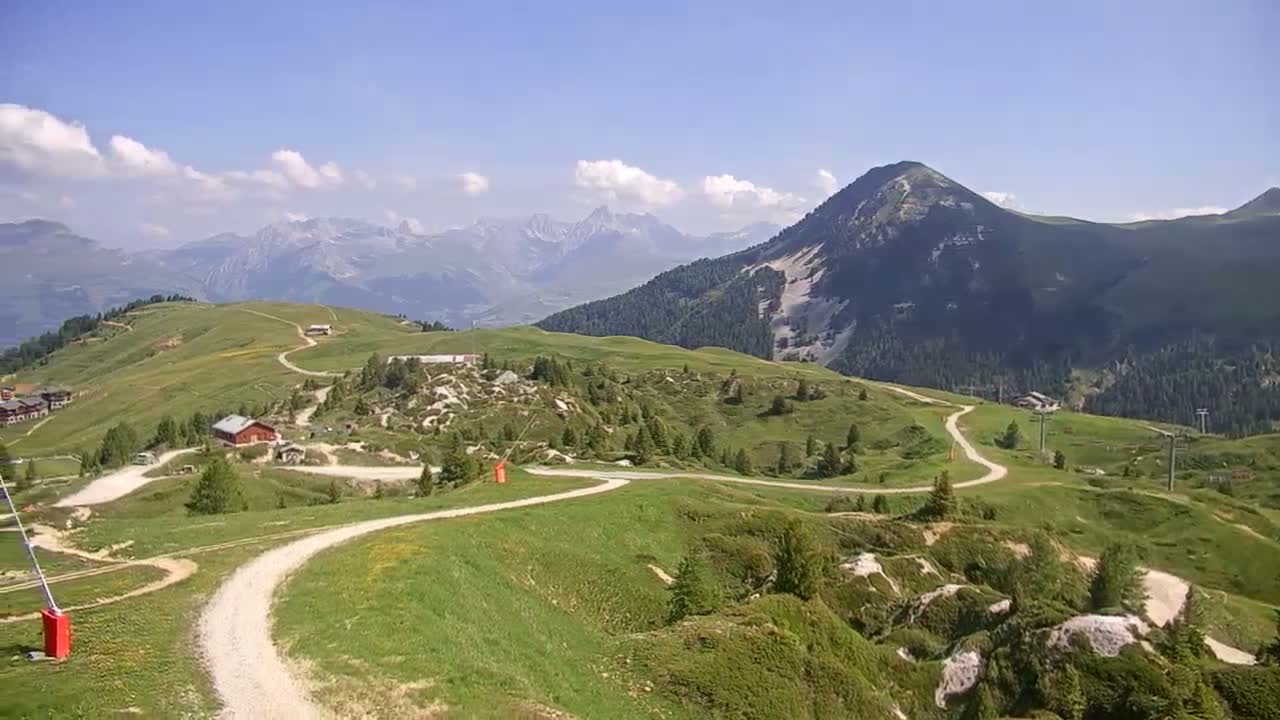 A winding dirt road traverses a grassy mountainside, with a few buildings visible, under a mostly sunny sky and a backdrop of distant mountains.