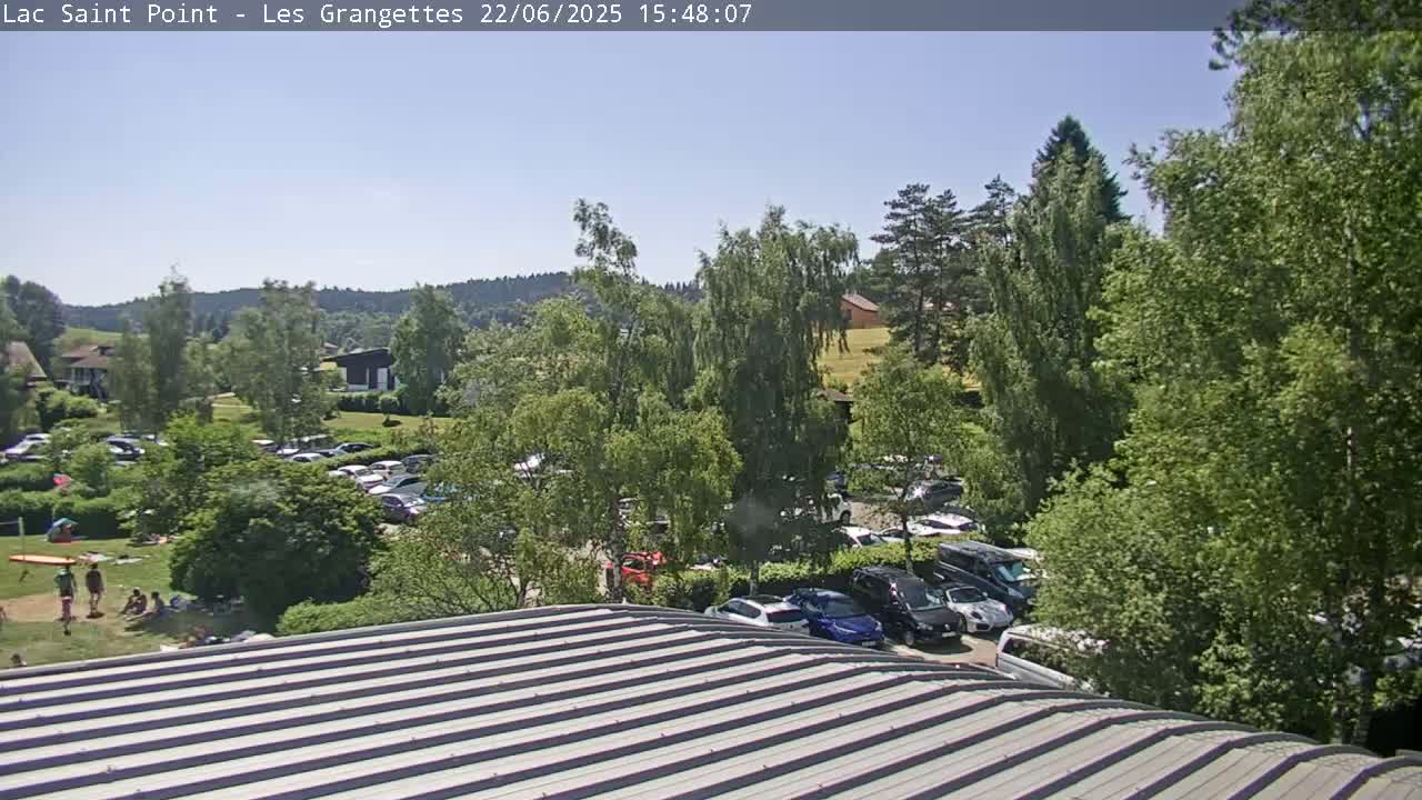 A view from a ridged roof shows a sunny day with a full parking lot, green trees, and people relaxing in a grassy area near buildings in the distance.