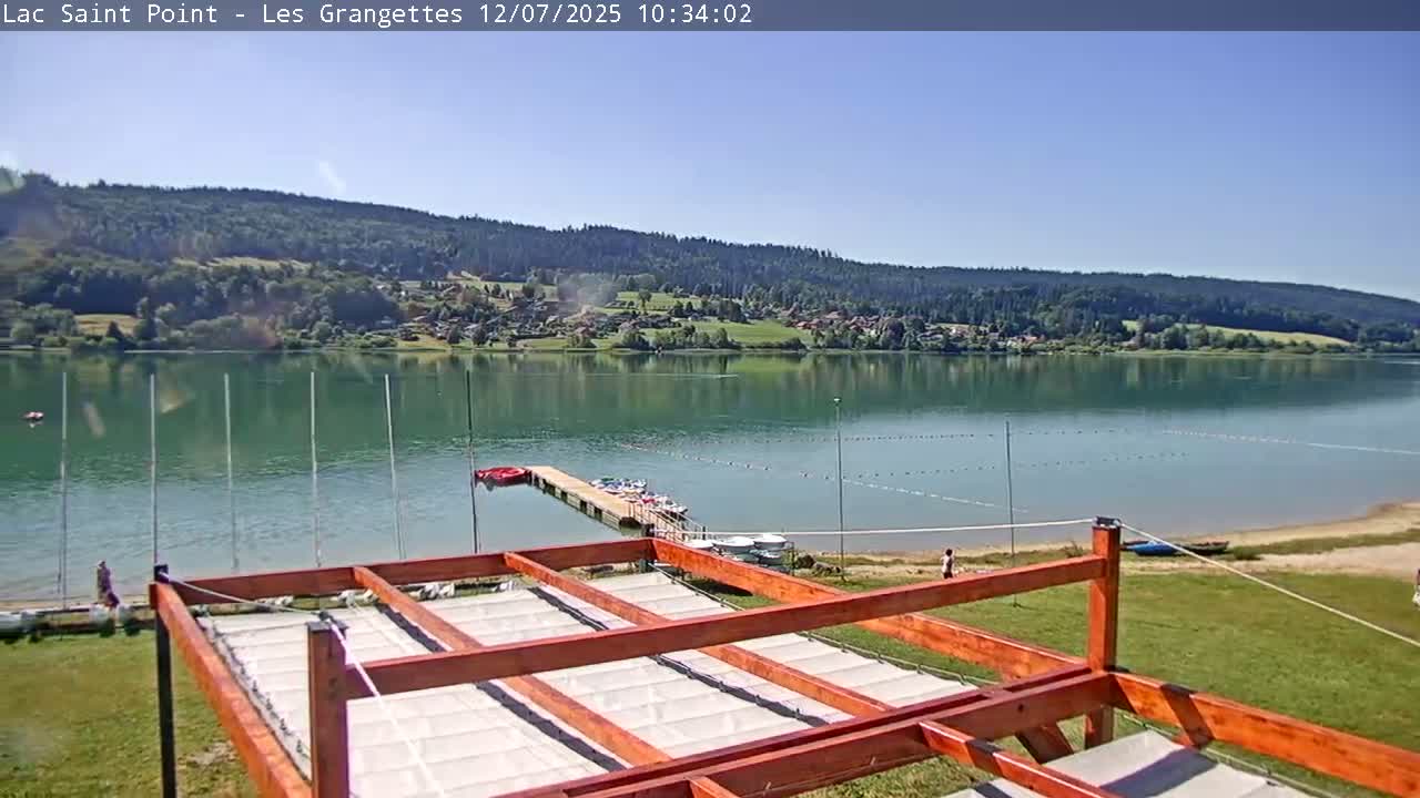 A calm lake stretches before a wooden structure with fabric-covered panels, overlooked by a verdant hillside under a clear, sunny sky.