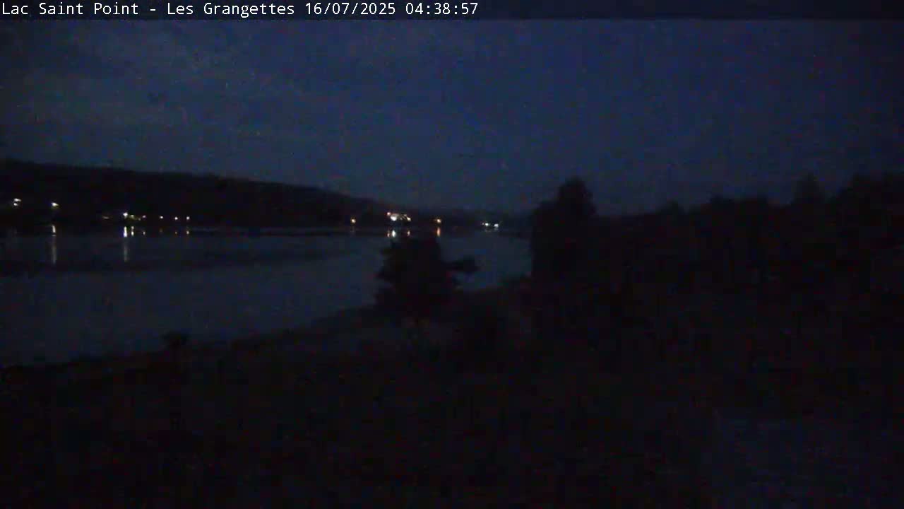A dark, nighttime scene shows a calm body of water reflecting a few distant lights, with silhouetted figures near the water's edge.