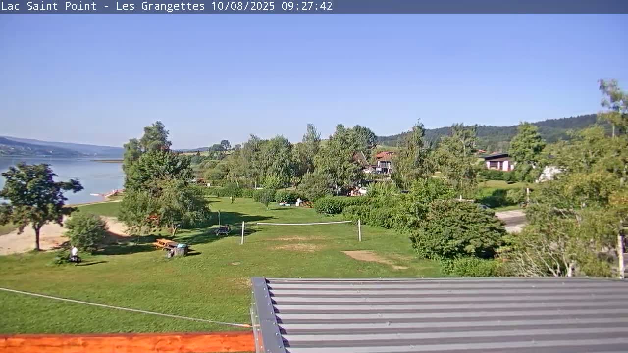 A sunny view overlooking a grassy area with a volleyball net near a lake and houses, under a clear blue sky.