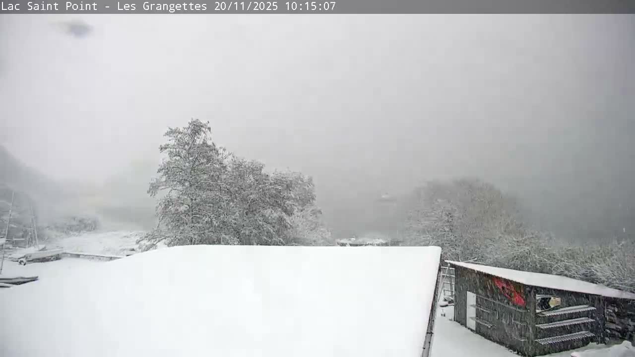 A wintry landscape is heavily blanketed in snow, featuring snow-laden trees, boats docked near a partially visible body of water, and a rustic wooden shed, all under an overcast and foggy sky with ongoing or recent heavy snowfall.