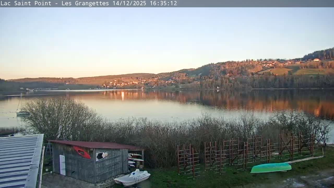 A wintry landscape is heavily blanketed in snow, featuring snow-laden trees, boats docked near a partially visible body of water, and a rustic wooden shed, all under an overcast and foggy sky with ongoing or recent heavy snowfall.