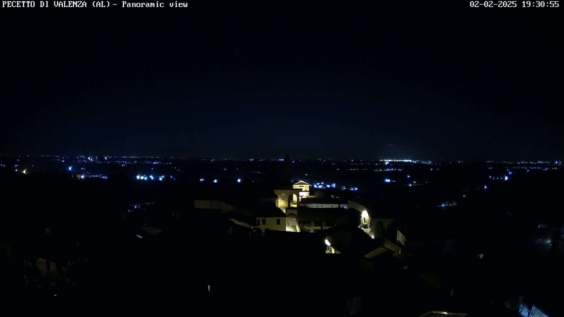 A nighttime panoramic view shows a small town illuminated with scattered lights, overlooking a larger, more distant city skyline under a clear, dark sky.