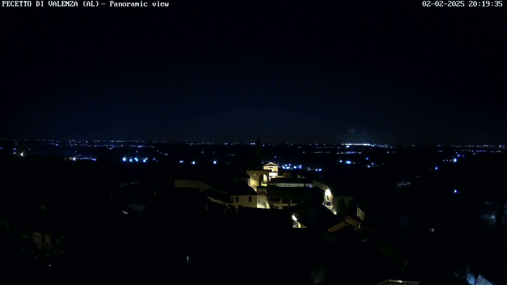 A nighttime panoramic view of a small town illuminated by scattered lights, with a distant city skyline visible under a clear, dark sky.