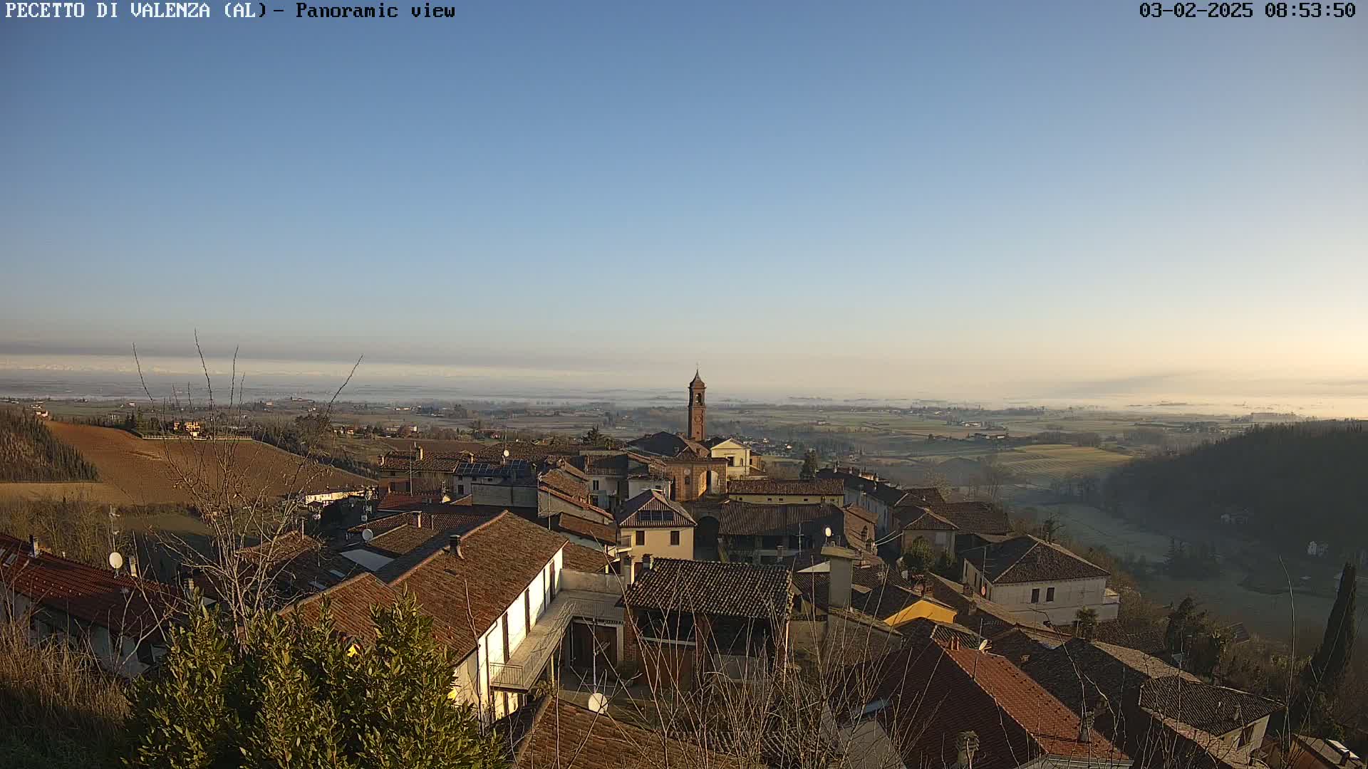 A panoramic view of a village nestled in a valley on a clear, slightly foggy morning, with rolling hills and farmland stretching into the distance.