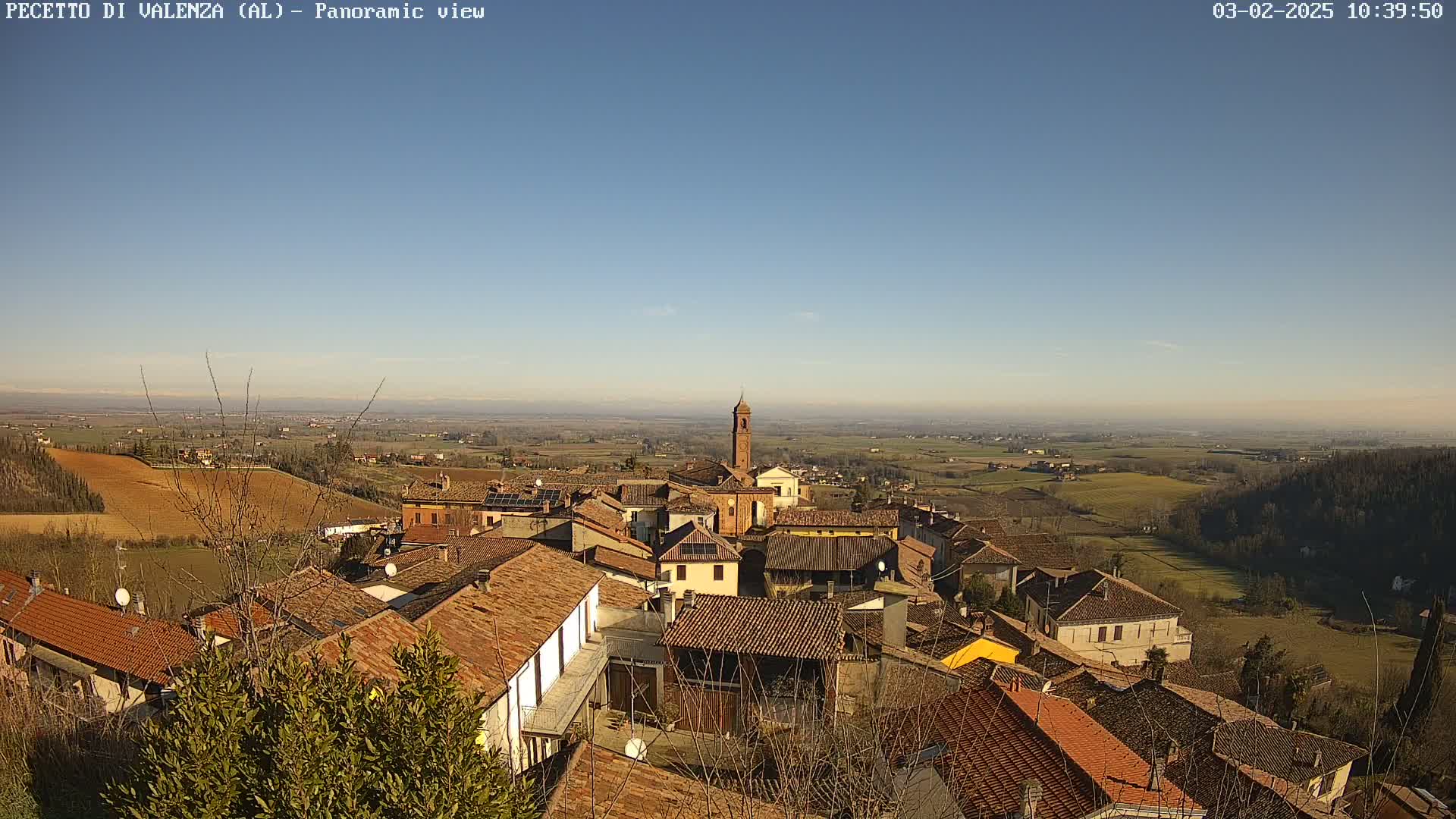 A panoramic view of a small town with terracotta-tiled roofs, nestled in a gently rolling landscape under a clear blue sky.