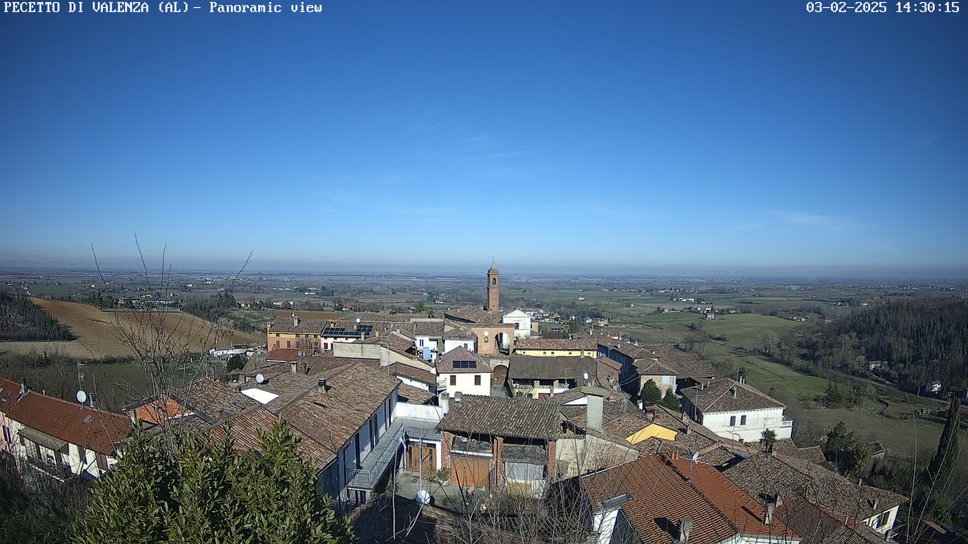 A panoramic view of a small town nestled in a valley under a clear blue sky, surrounded by gently rolling hills and farmland.