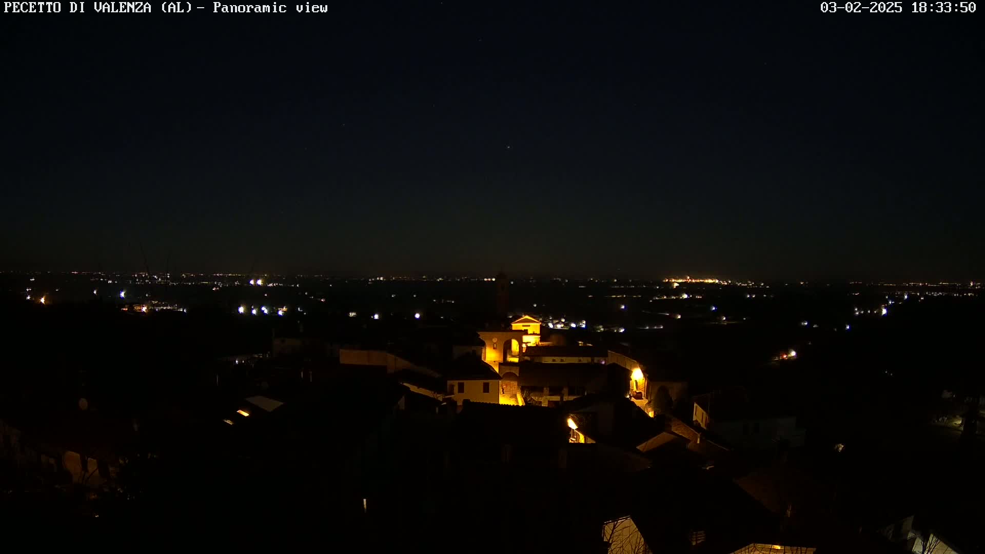 A nighttime panoramic view shows a small, illuminated town nestled in a dark landscape, with a distant city skyline visible under a clear, starlit sky.