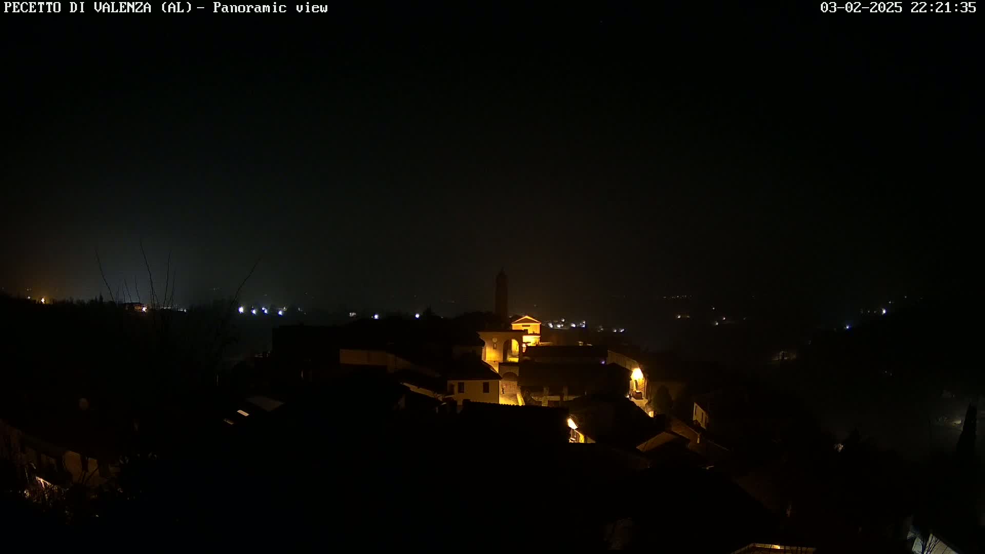 A nighttime panoramic view of a small town nestled in a valley, dimly lit with a few scattered lights and shrouded in a hazy atmosphere.
