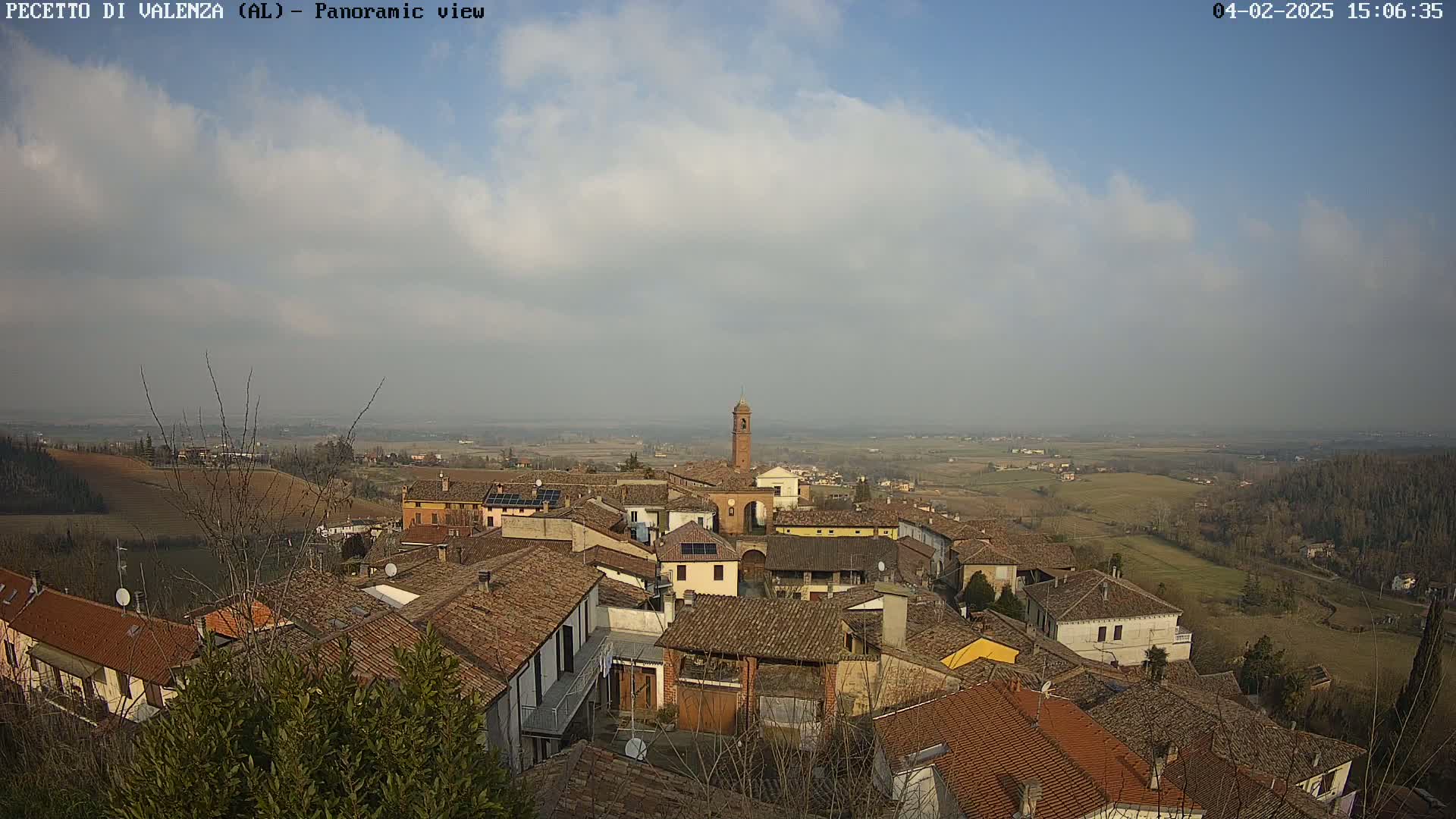 A panoramic view shows a village of red-tiled roofs nestled in a gently rolling landscape under a partly cloudy sky.