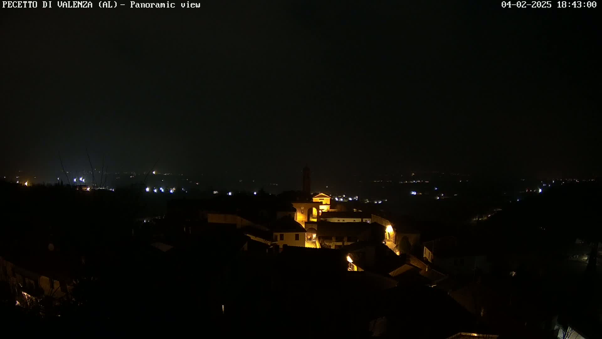 A nighttime panoramic view of a small, illuminated town nestled in a dark, hilly landscape under a clear, dark sky.