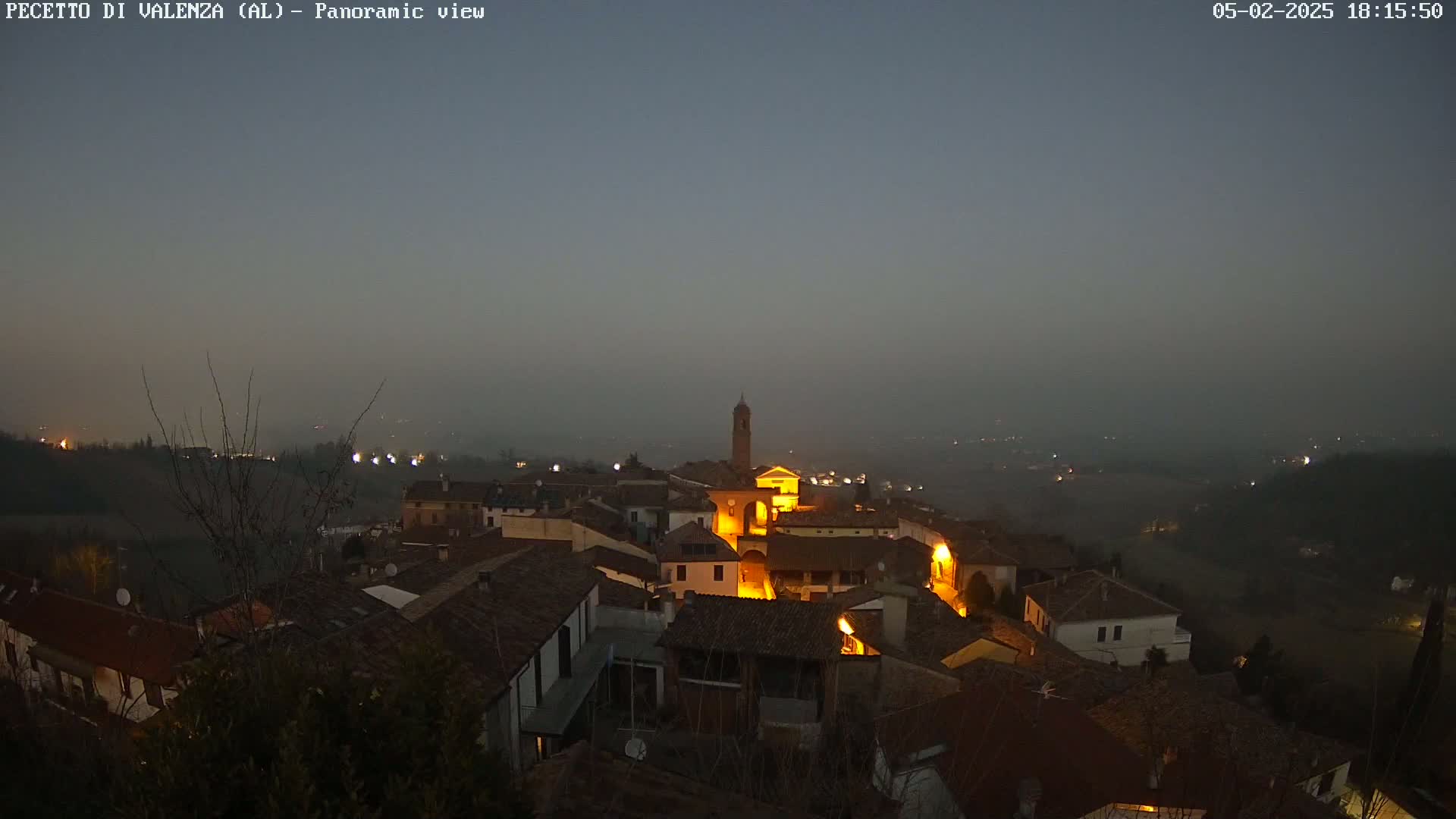 A hazy, twilight view of a small, illuminated village nestled in a valley, with a church tower prominent in the center.
