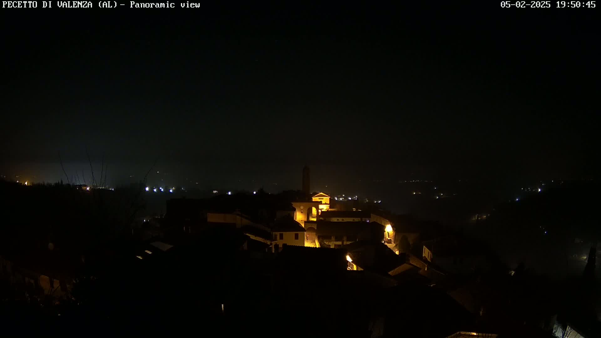 A nighttime panoramic view of a small, dimly lit town nestled on a hill, under a dark, possibly hazy sky.