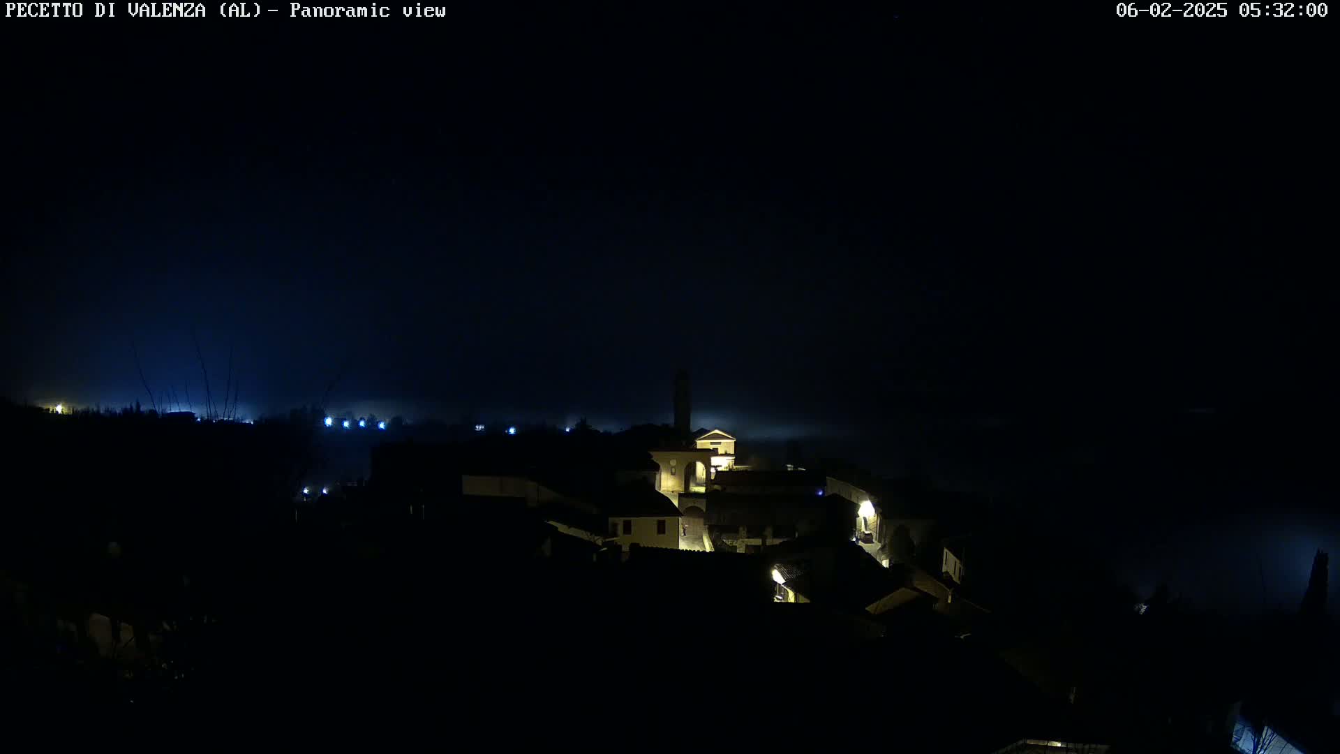A nighttime panoramic view shows a small, dimly lit town shrouded in fog or mist under a dark sky.