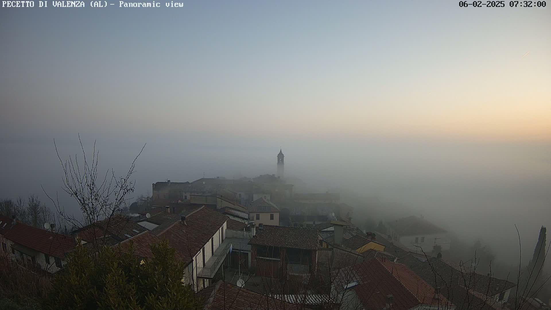A foggy sunrise reveals a village of red-tiled rooftops and a bell tower, partially obscured by mist.