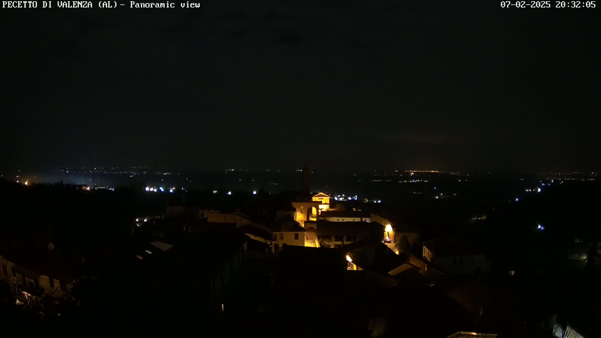 A nighttime panoramic view shows a small, illuminated town nestled among dark hills, with a distant horizon speckled with more lights.