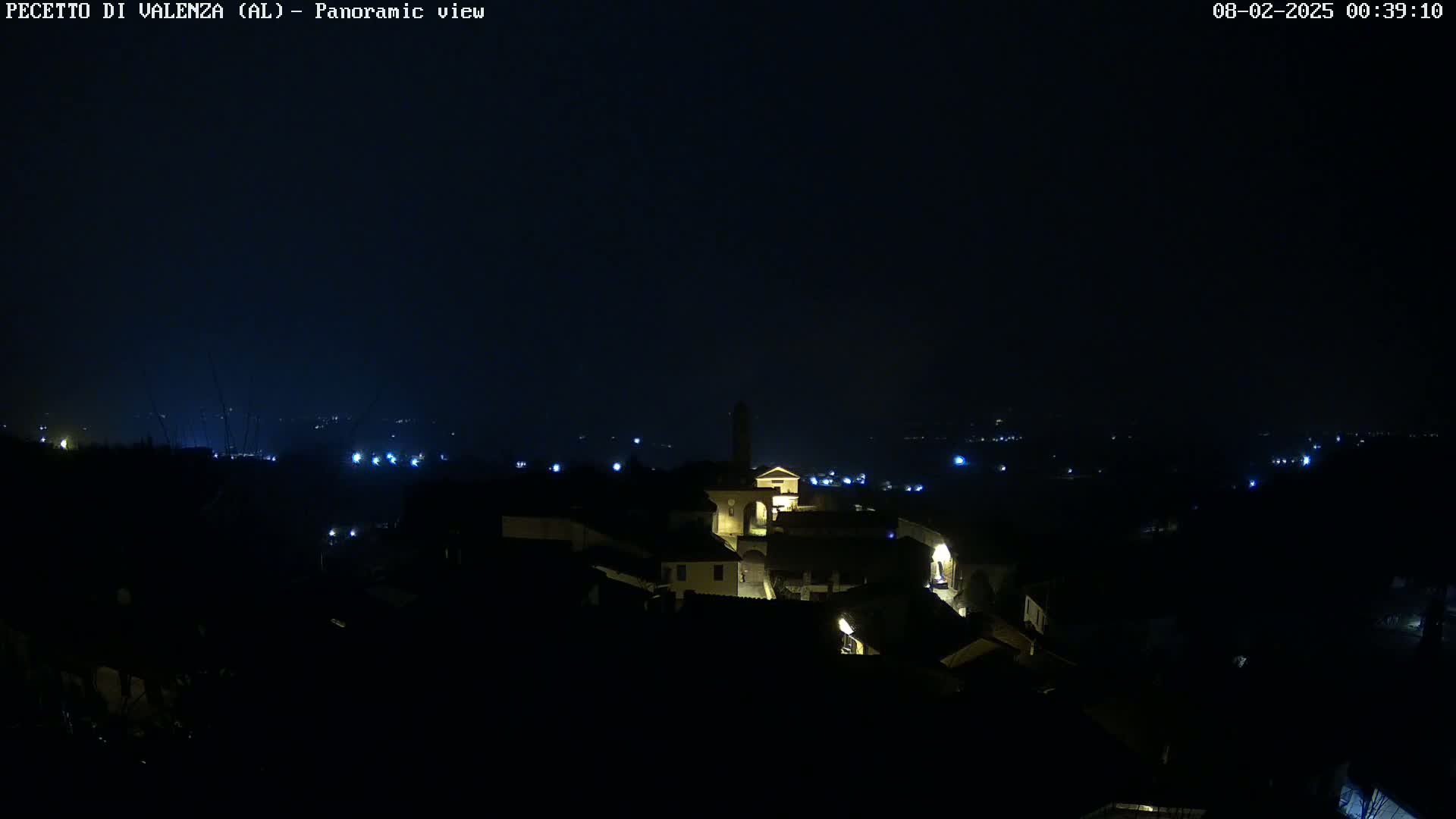 A nighttime panoramic view of a small, dimly lit town with a few illuminated buildings, under a dark, clear sky.