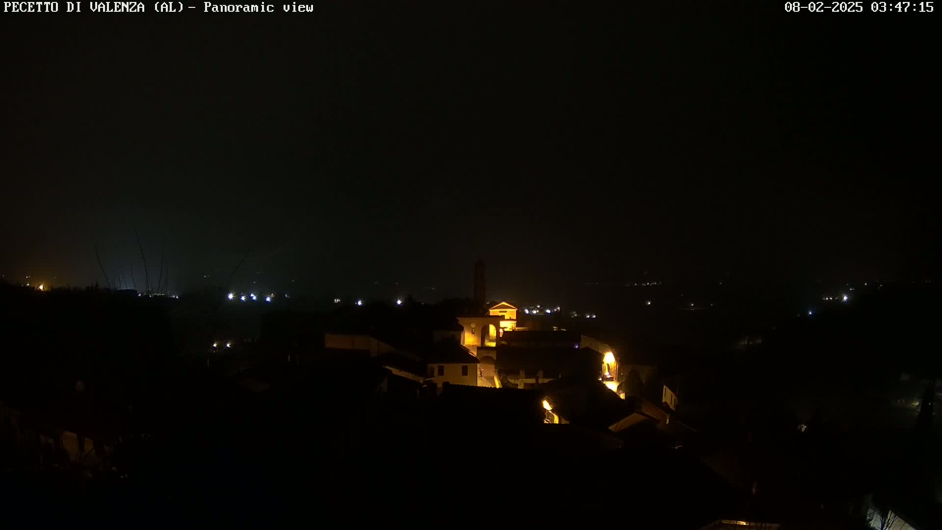 A nighttime panoramic view shows a small, dimly lit town nestled in a dark landscape under a hazy, dark sky.