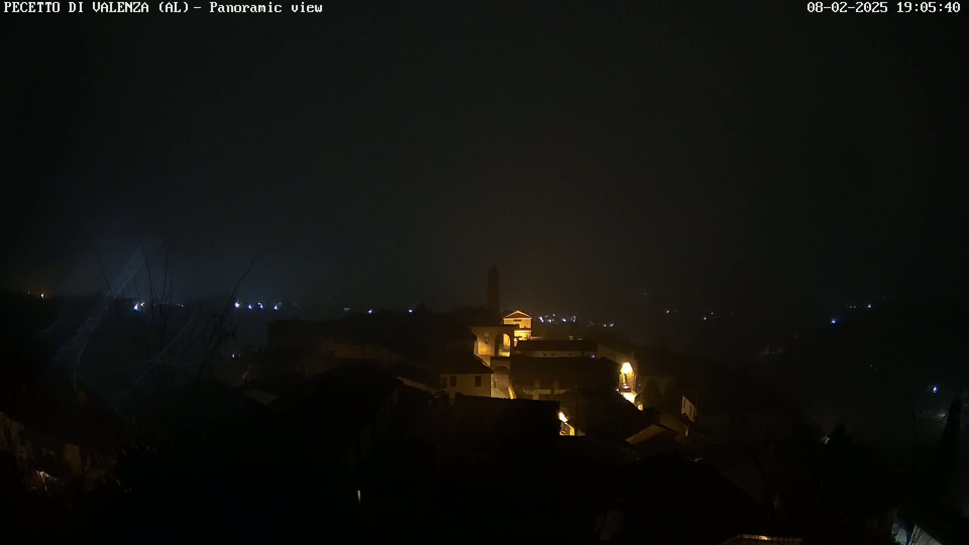 A nighttime panoramic view of a small town shrouded in fog, with dimly lit buildings and a church tower visible in the center.