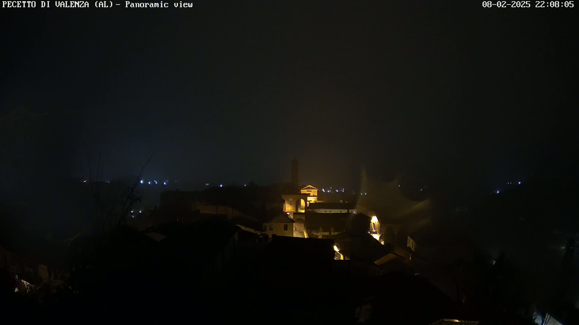 A nighttime panoramic view of a small town nestled in a foggy or misty landscape, with a few illuminated buildings visible.