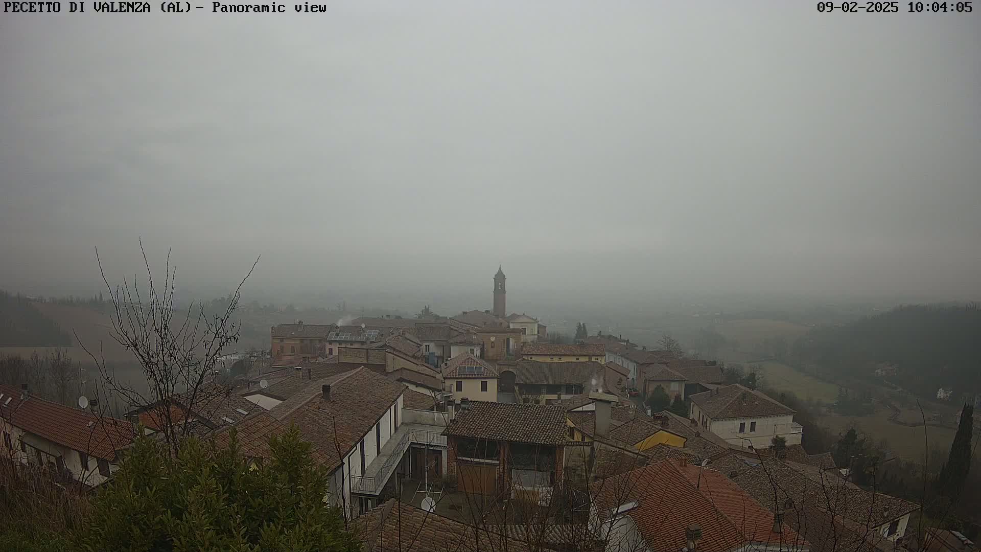 A foggy, panoramic view of a small town nestled in a valley, with rooftops of mostly brown tile and a tall bell tower visible in the center.