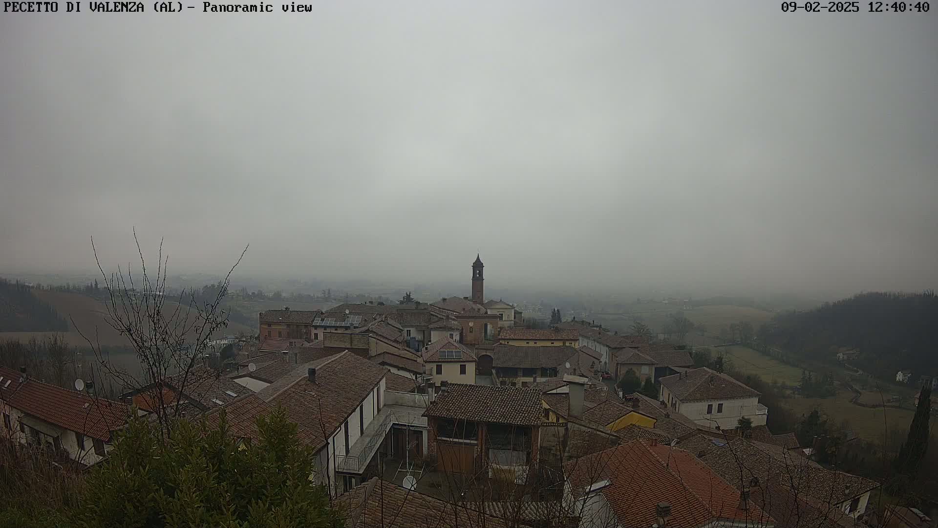 A foggy view of a small town nestled in a valley, with numerous brown-roofed buildings and a tall bell tower visible in the center.