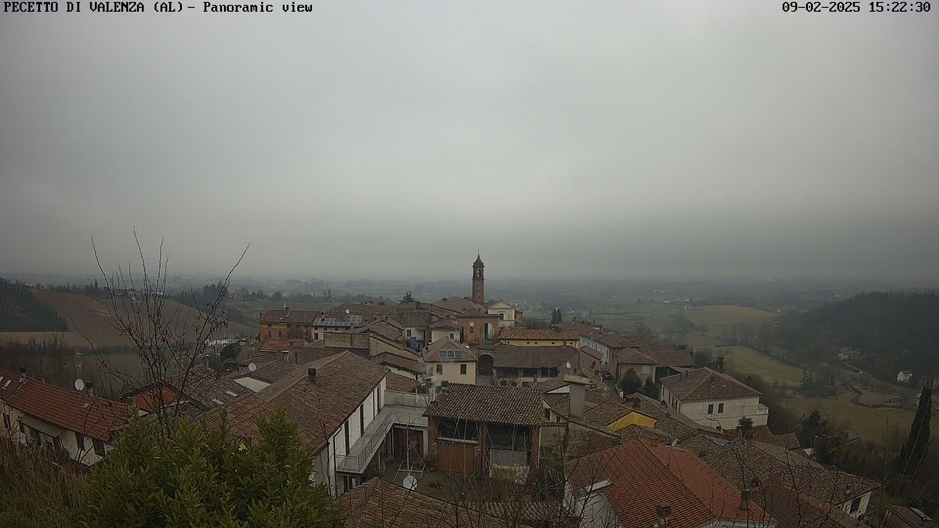 A panoramic view of a village with brown-tiled rooftops and a bell tower, under a hazy, overcast sky.