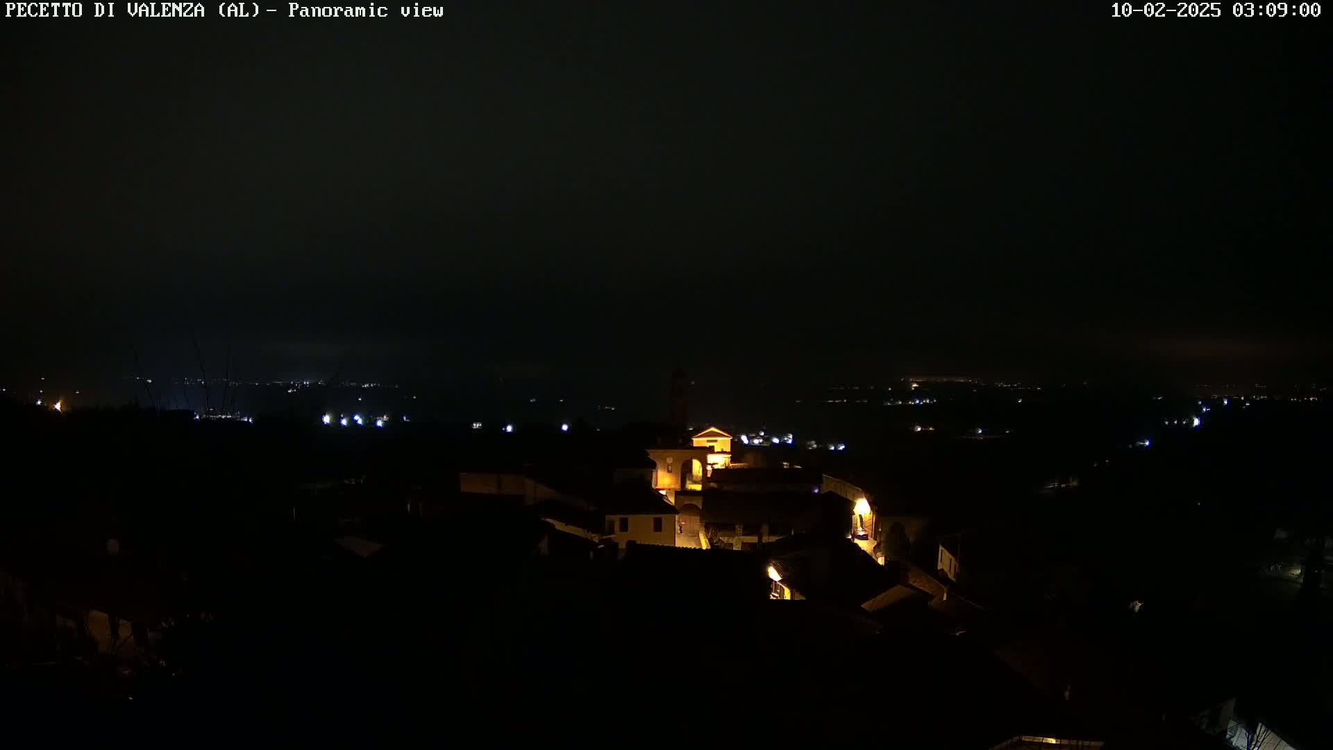 A nighttime panoramic view shows a small, illuminated town nestled in a dark, hilly landscape under a cloudy sky.