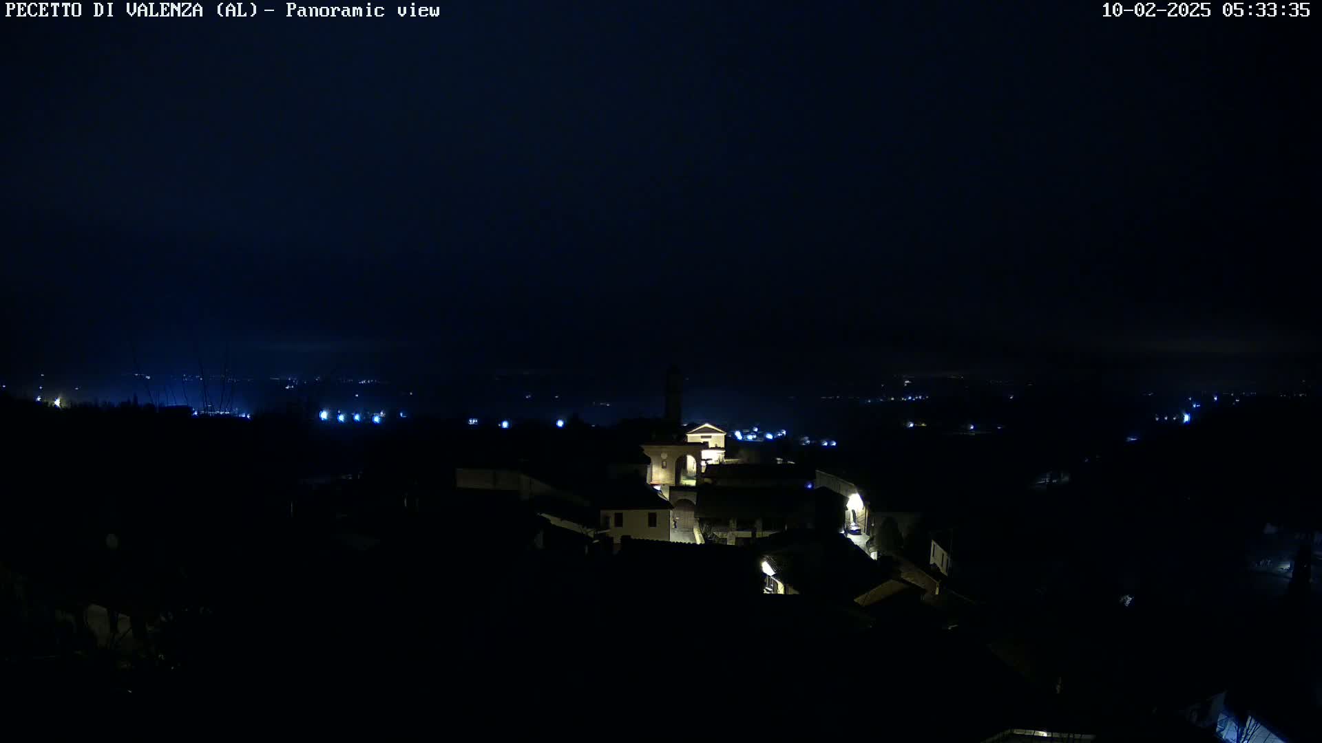 A dark, nighttime panoramic view of a small, illuminated town nestled in a valley under a clear, dark sky.
