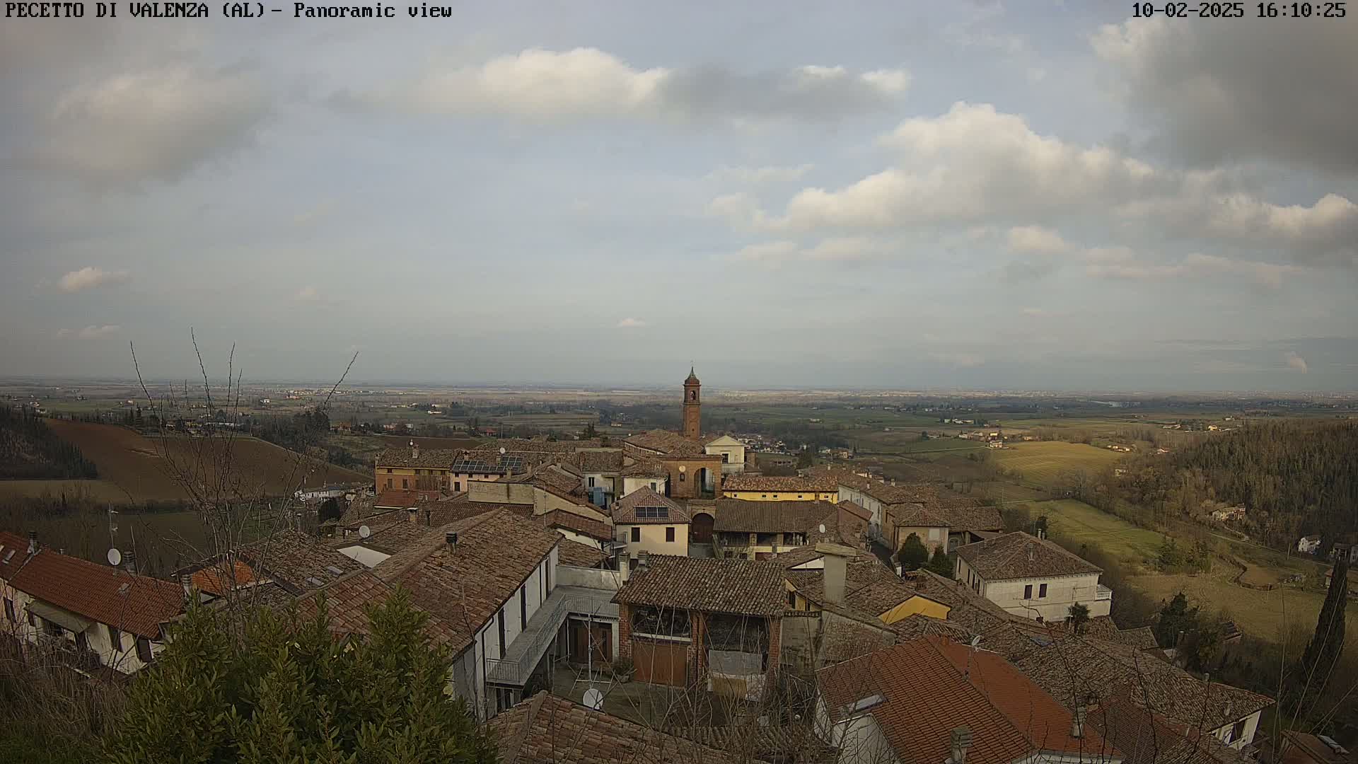 A panoramic view of a small town with terracotta roofs, nestled in a hilly landscape under a partly cloudy sky.