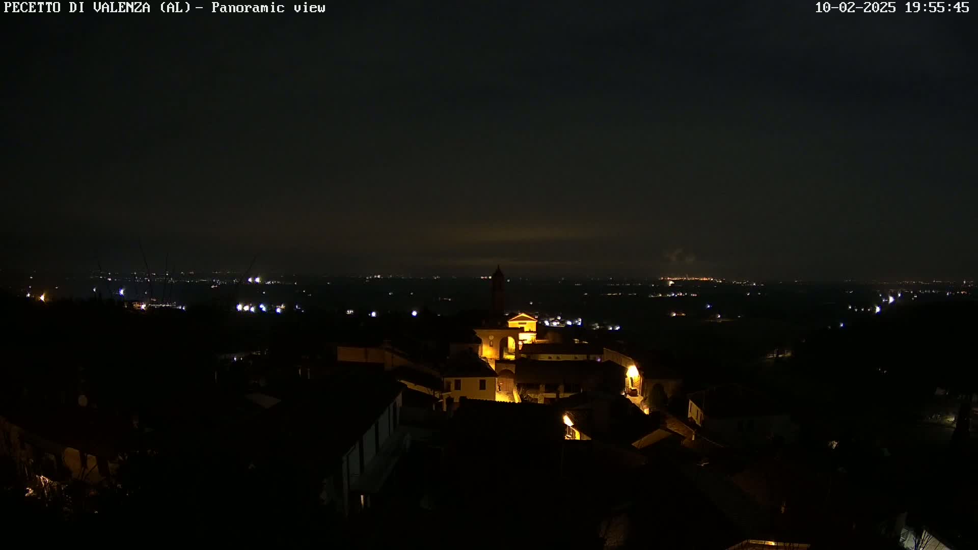 A nighttime panoramic view shows a small, illuminated village nestled in a dark landscape, with distant city lights visible under a mostly clear, dark sky.