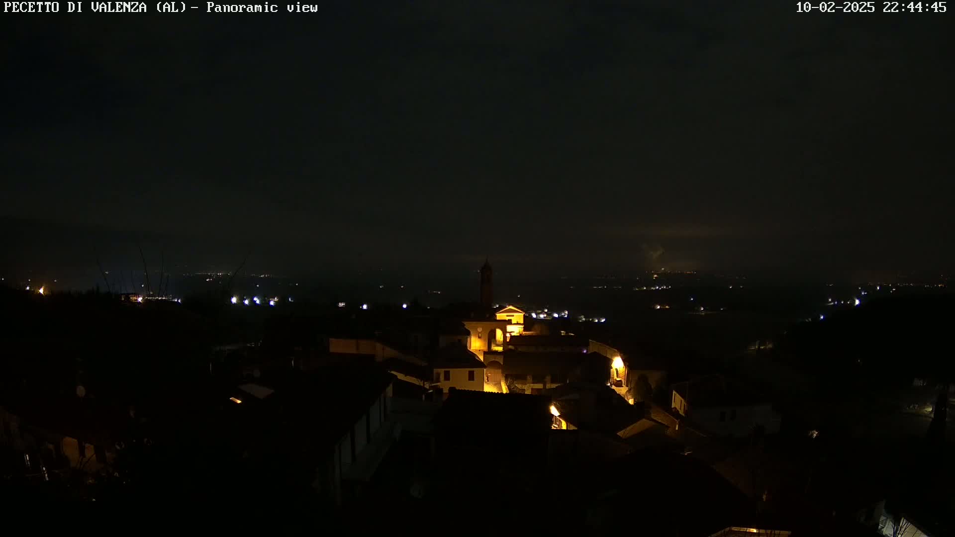 A nighttime panoramic view of a small village, illuminated by scattered interior lights, under a dark, cloudy sky.