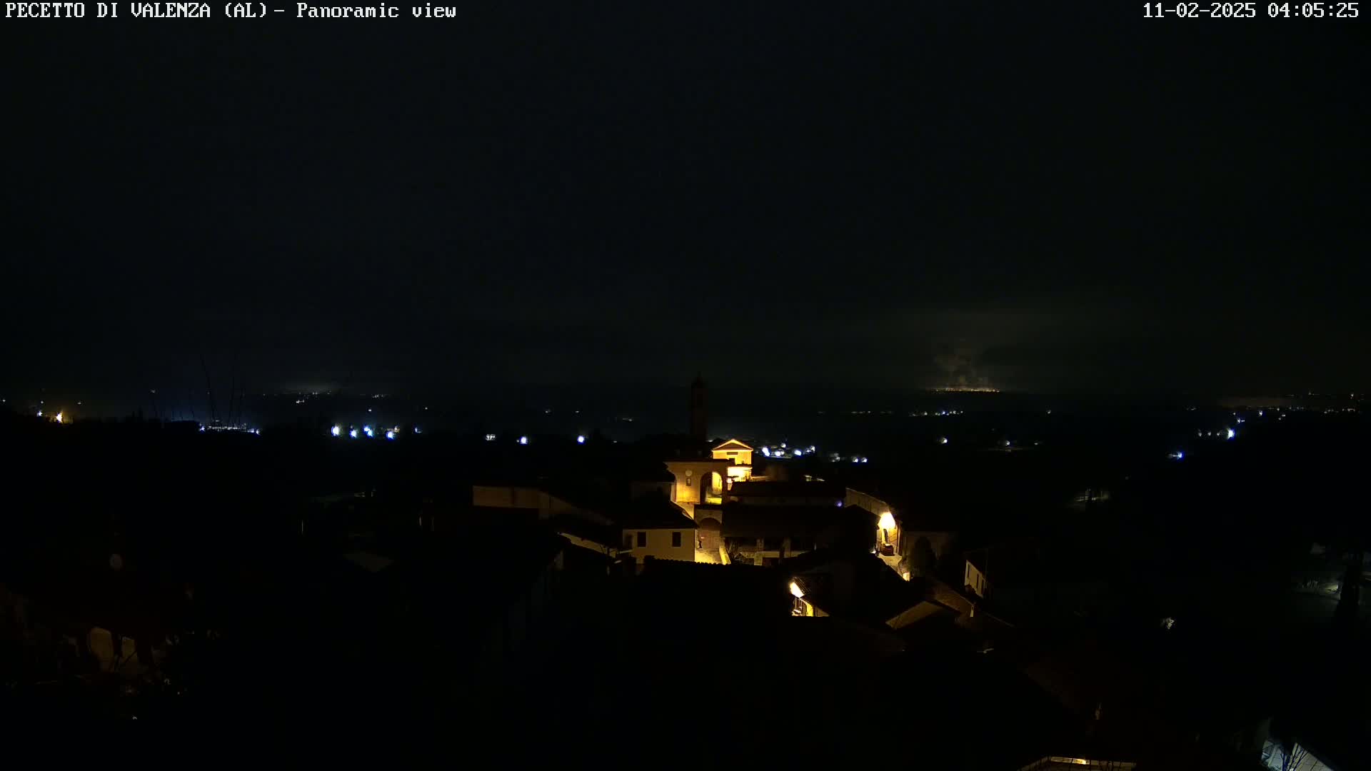 A nighttime panoramic view of a small, dimly lit town nestled in a dark landscape, with a distant glow possibly from another settlement or light pollution on the horizon.