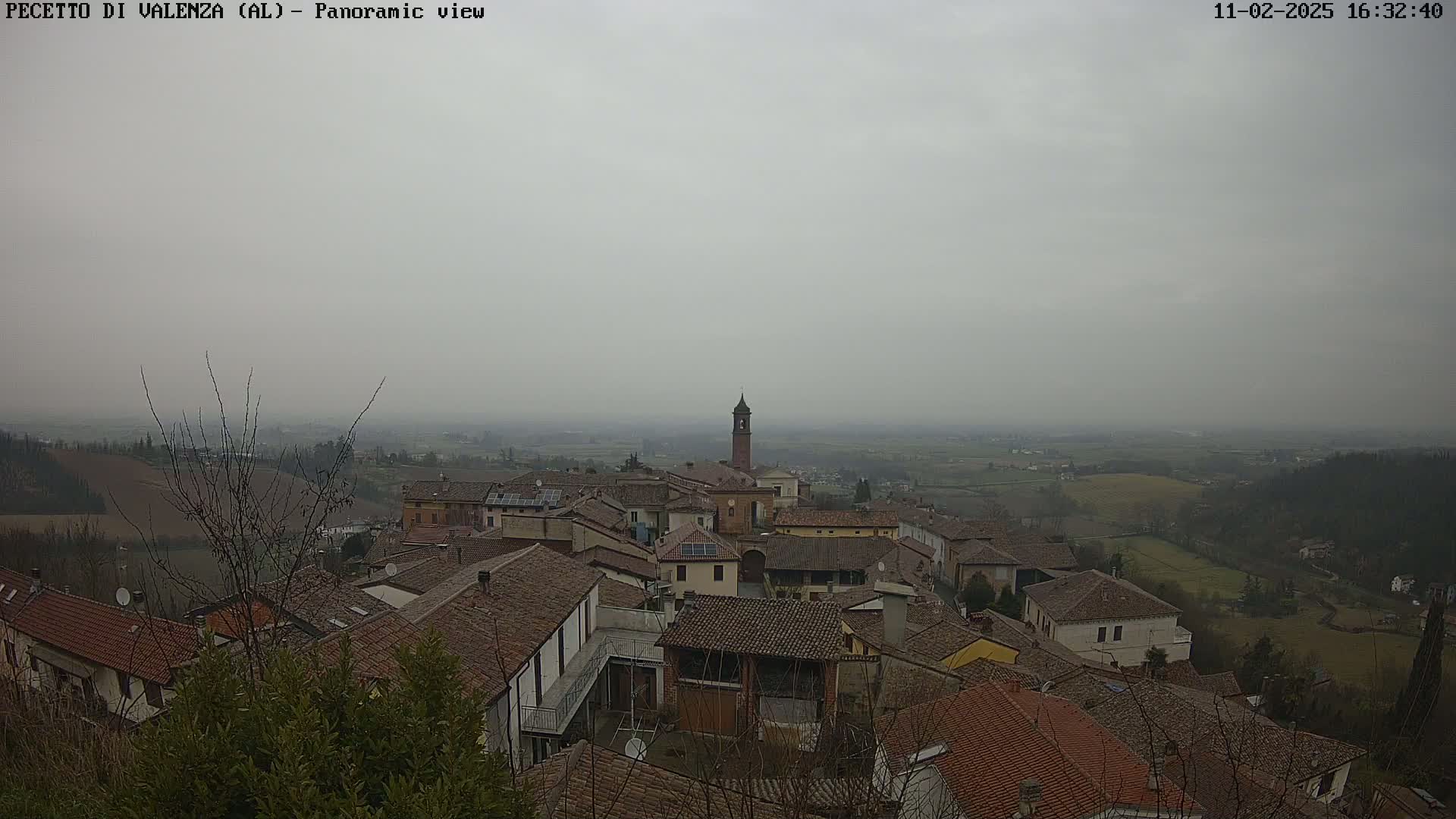 A hazy, overcast day reveals a panoramic view of a village nestled in a valley, with red-tiled roofs and a tall bell tower visible among the buildings.