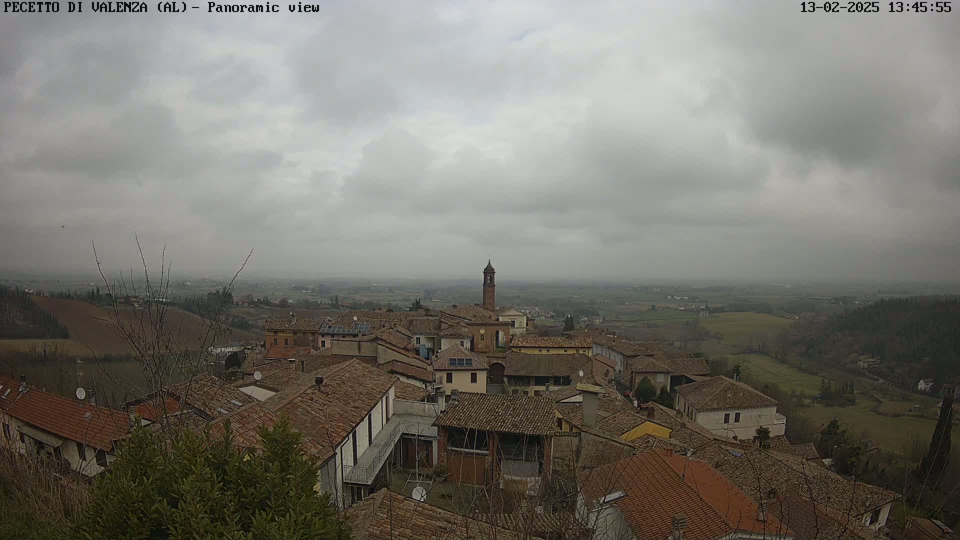 A panoramic view of a town with terracotta rooftops under an overcast sky shows a bell tower in the center.