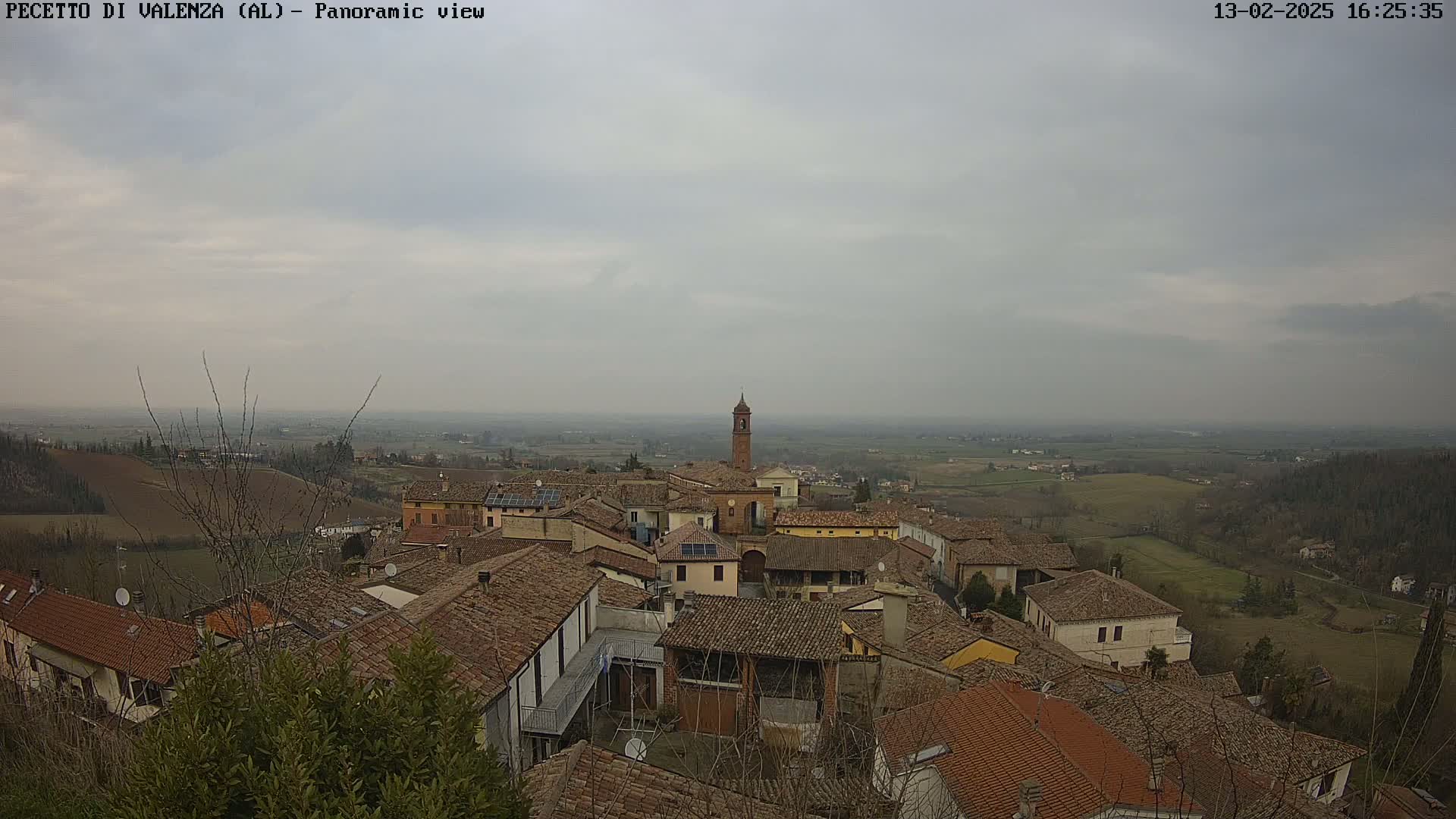 A panoramic view of a town with red-tiled roofs and a tall bell tower, under an overcast sky.