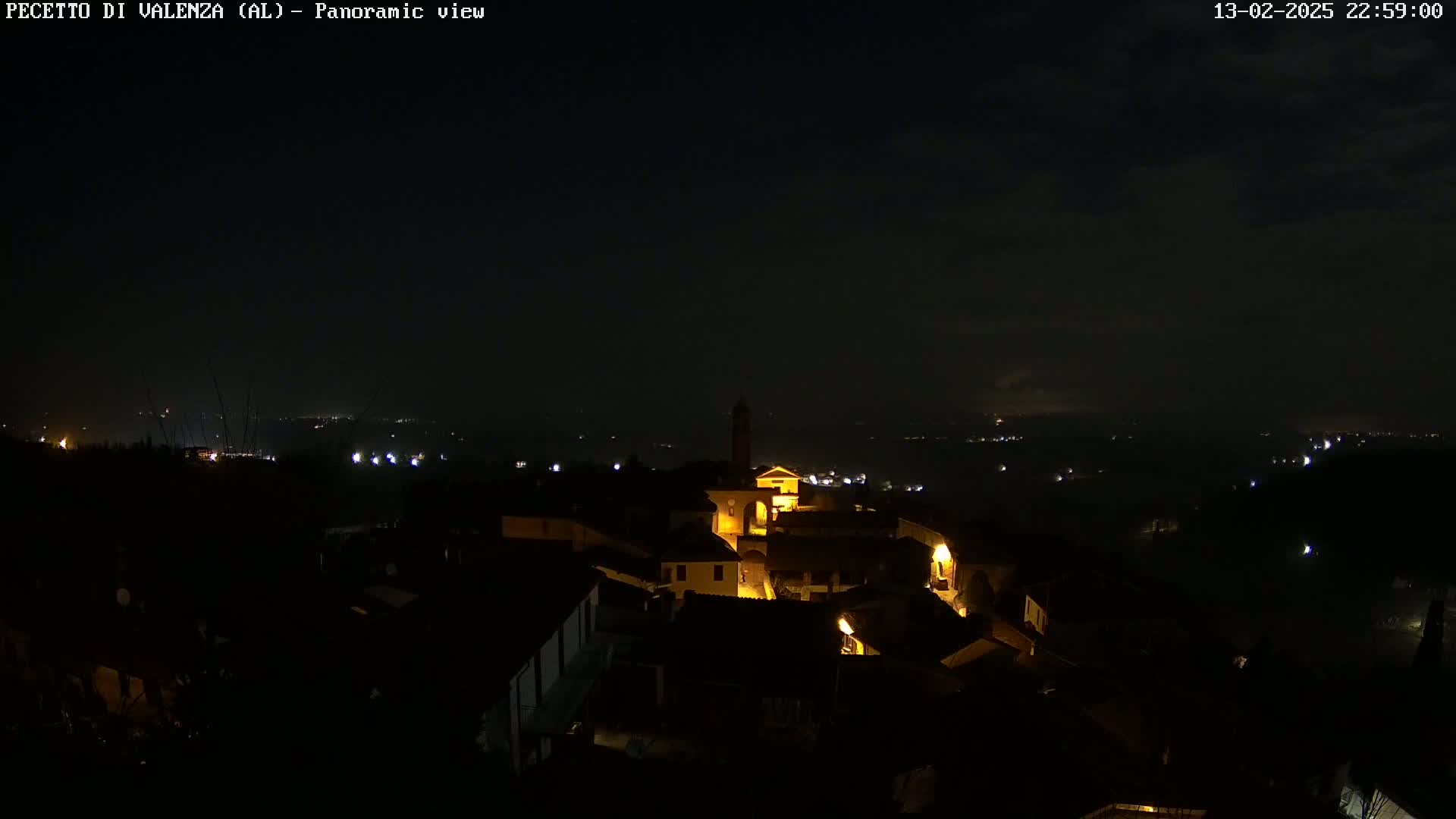 A nighttime panoramic view of a small, illuminated town nestled in a dark landscape under a partly cloudy sky.