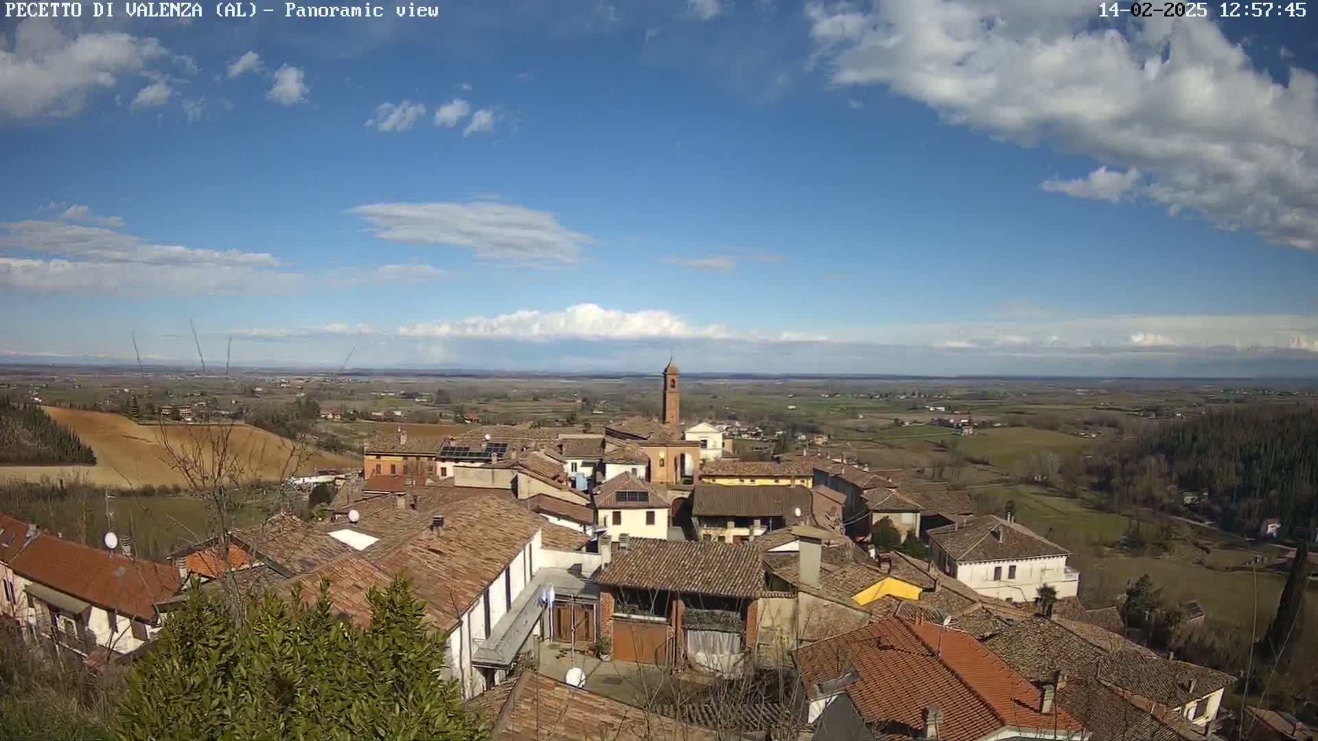 A panoramic view of a small town with terracotta rooftops, under a mostly sunny sky with scattered clouds.