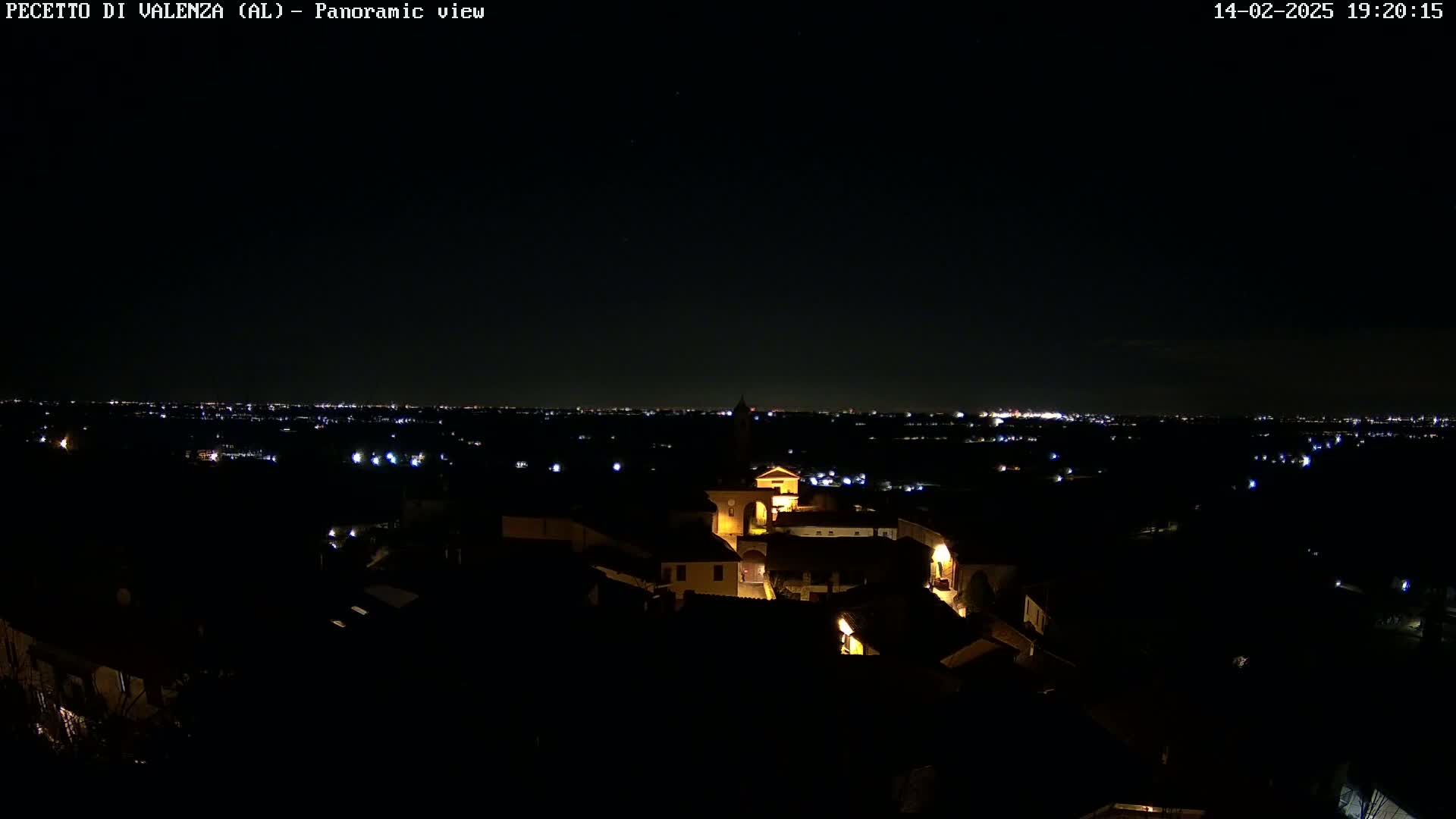 A nighttime panoramic view shows a small, illuminated village nestled in the dark landscape, with a distant city skyline glowing faintly on the horizon under a clear, dark sky.