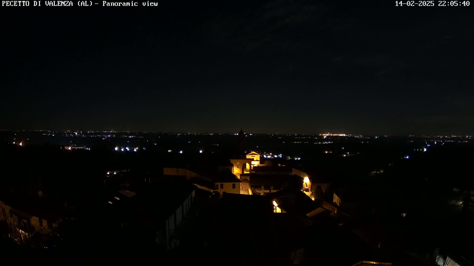 A nighttime panoramic view shows a small, illuminated town nestled amongst dark hills, overlooking a distant city skyline under a clear, dark sky.