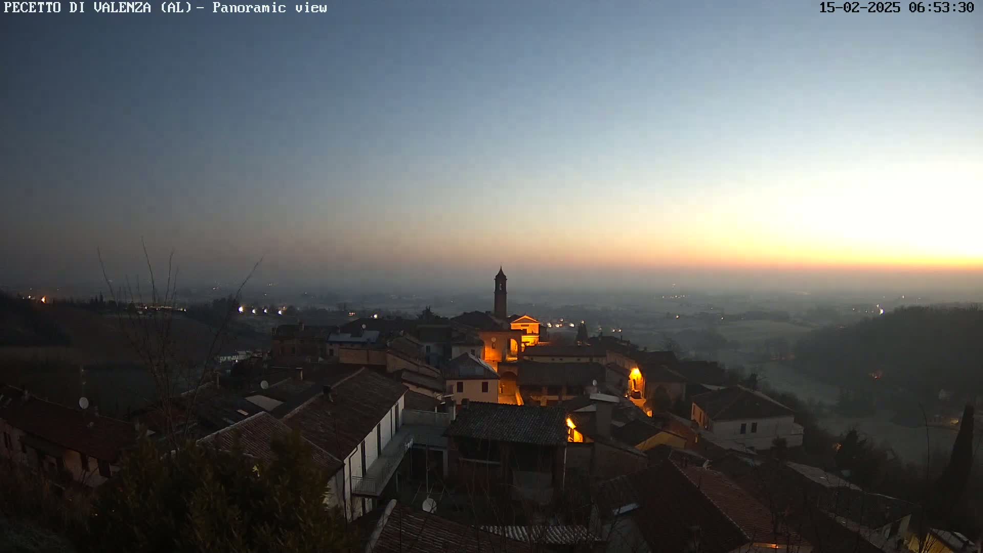 A hazy sunrise illuminates a small town nestled on a hill, overlooking a vast, flat landscape.