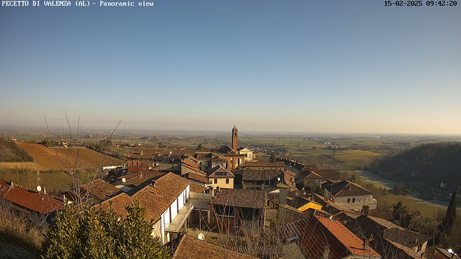 A panoramic view of a small town nestled in a hilly landscape on a clear, sunny day, with a church steeple prominent in the center.